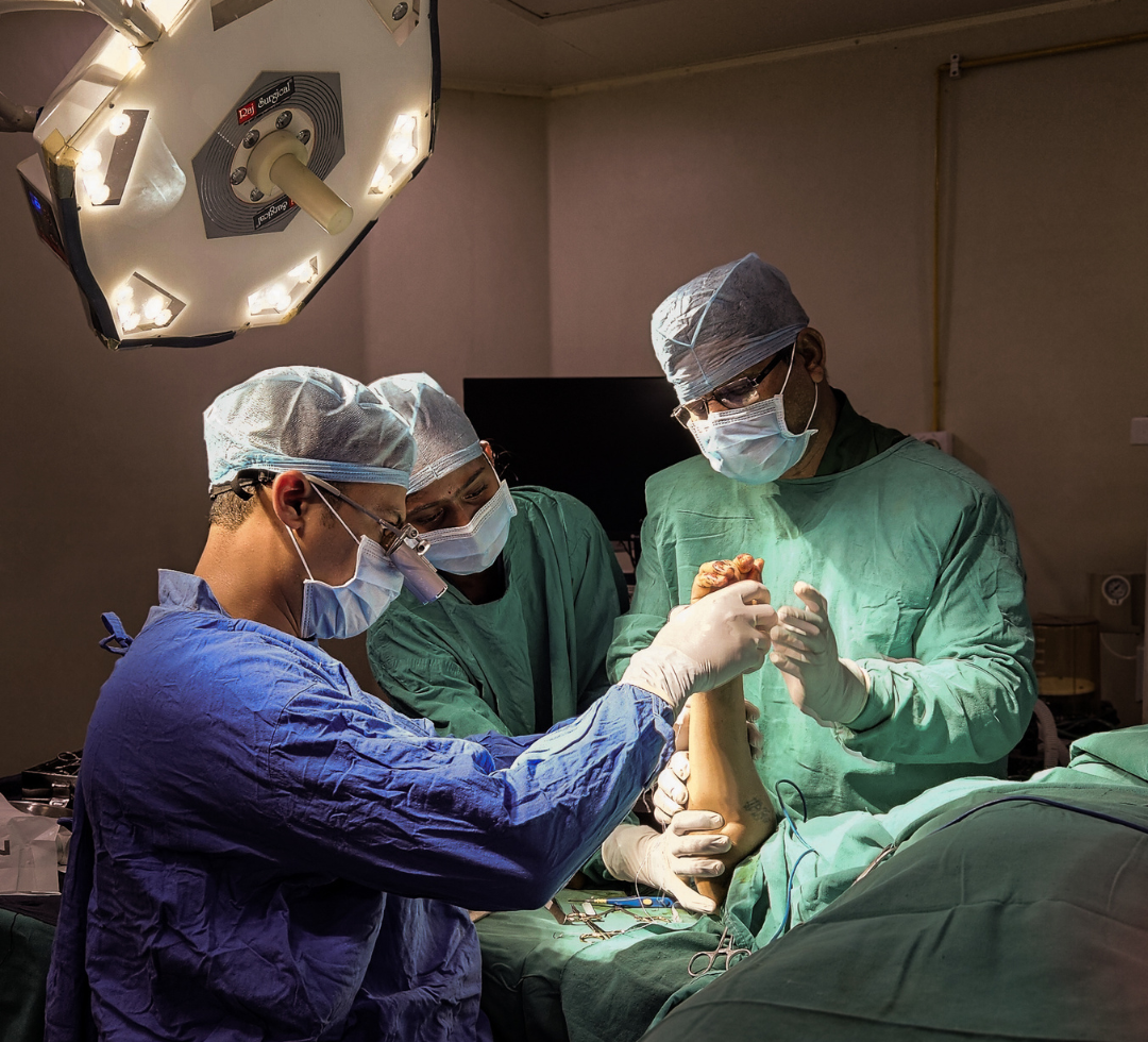 A surgeon in a sterile operating room performing a hand surgery procedure under bright surgical lights, assisted by a medical professional.